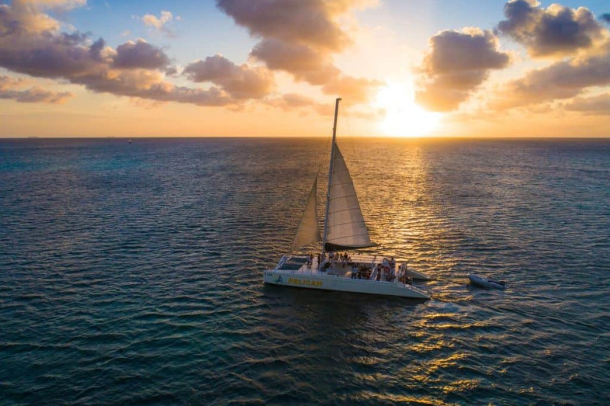 A Sunset Cruise (Aruba) sails smoothly across calm ocean waters at sunset, with people on board enjoying golden light that filters through scattered clouds on the horizon.