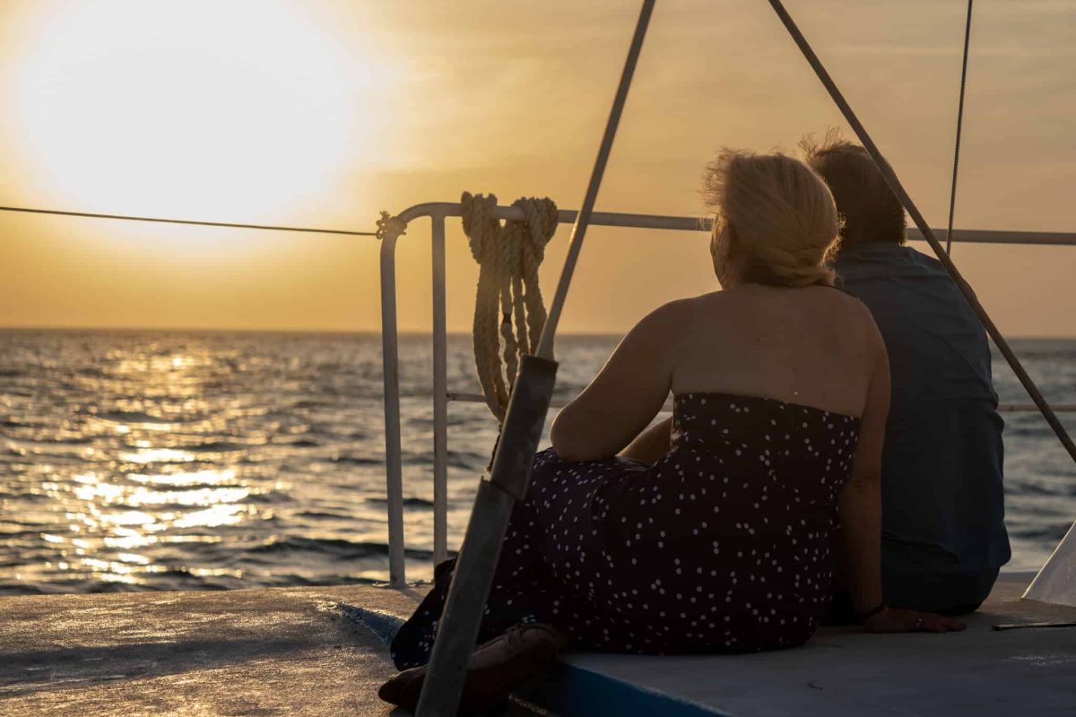 Two people relax side by side on a boat during the Sunset Cruise (Aruba), watching the sun dip below a tranquil ocean. Ropes and railings frame the view as golden light glimmers off the water, creating a serene, romantic mood.