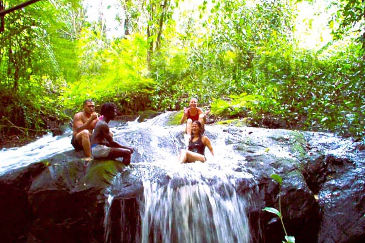 Vier mensen relaxen op rotsen bij een kleine waterval in een weelderig groen bos en glimlachen terwijl het water onder hen stroomt. Ervaar deze natuurlijke schoonheid met de 4 Dagen Raleighvallen Voltzberg tour in Suriname.