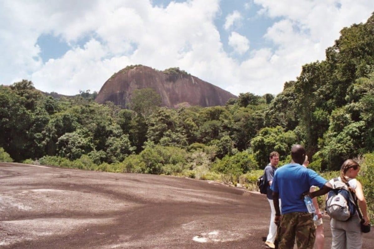 Drie wandelaars verkennen de Raleighvallen Voltzberg in Suriname, staand op een rotsachtige ondergrond temidden van weelderige bossen met de iconische granieten berg op de achtergrond onder een gedeeltelijk bewolkte hemel.