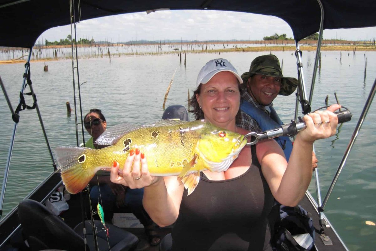 Tijdens de Vistour op de Monding Surinamerivier houdt een lachende vrouw op een boot een grote, kleurrijke vis vast terwijl twee mannen achter haar zitten op een meer met boomstronken en een gedeeltelijk bewolkte lucht.