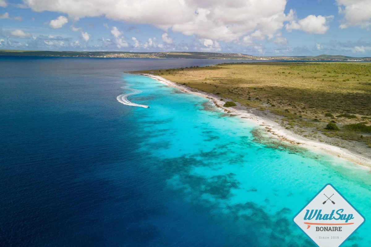 Vista aérea de una clara costa turquesa con un barco dejando un rastro blanco en el agua, bordeado por pastizales secos bajo un cielo parcialmente nublado. Un crucero privado de lujo con el logo del atardecer (Bonaire) aparece en la esquina inferior derecha.