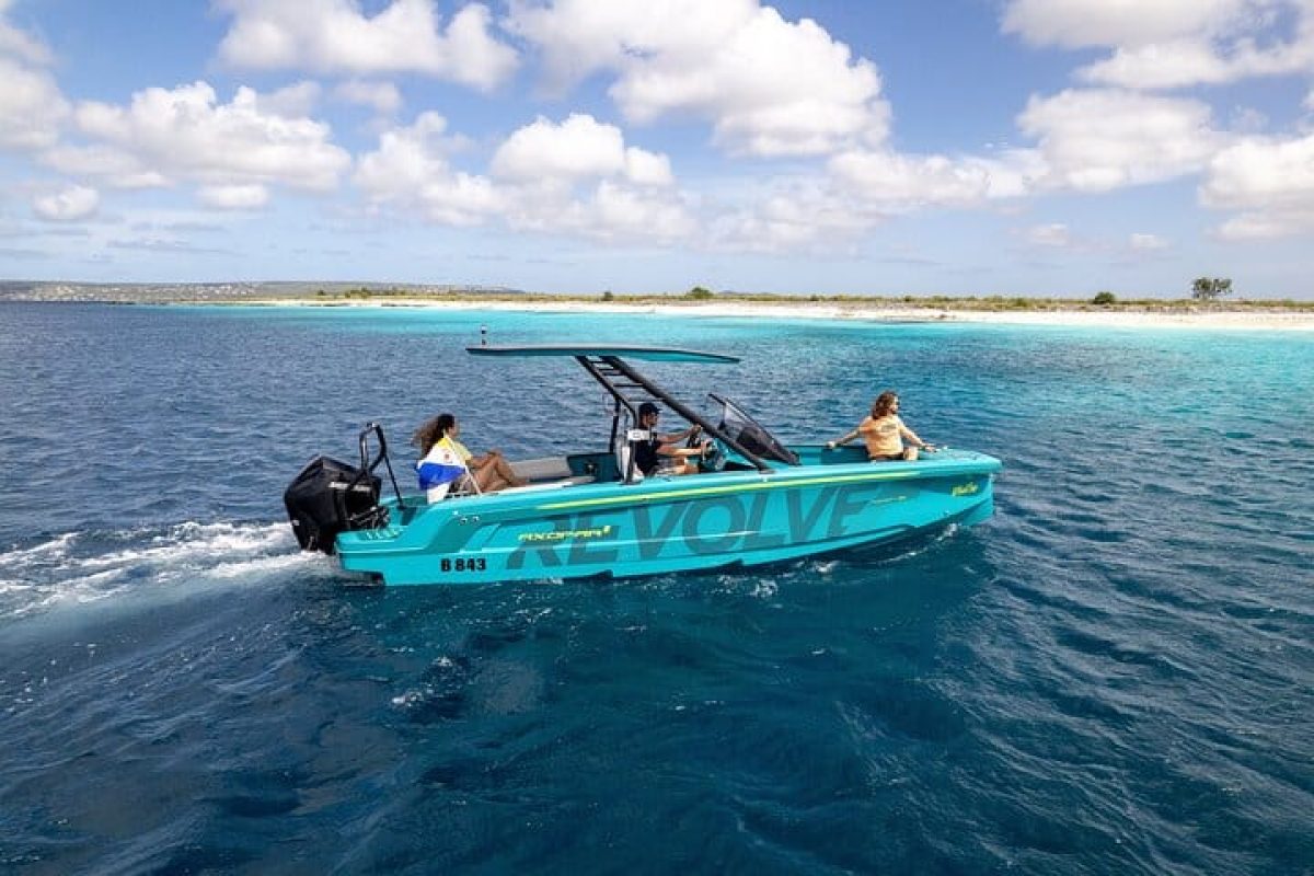 Un crucero privado de lujo al atardecer (Bonaire) navega con tres personas en aguas claras y azules a lo largo de una playa de arena bajo un cielo parcialmente nublado.