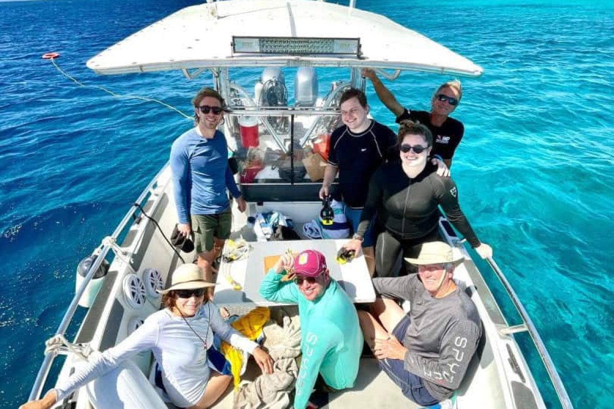 A group of seven people smiles and poses on a small boat during the 2 Tank Boat Dive (Bonaire), enjoying food and drinks over clear turquoise waters near a tropical island under a blue sky with scattered clouds.