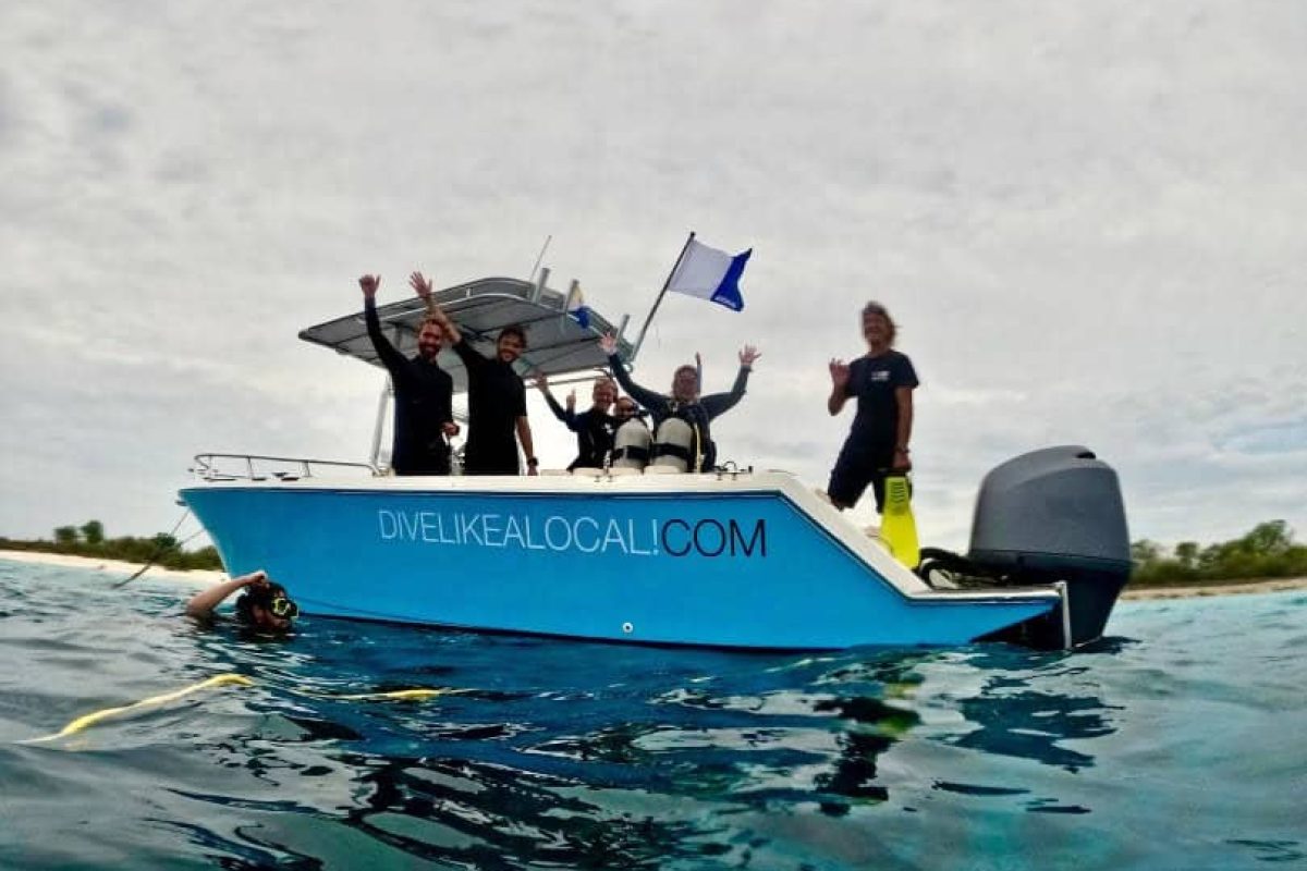 Un grupo disfruta de un paseo en barco con una parada para hacer snorkel (Bonaire) en un barco azul y blanco con DIVELIKEALOCALI.COM, saludando a la cámara mientras sostiene una bandera de buceo. El barco flota en aguas tranquilas bajo las nubes, con un buceador nadando cerca.