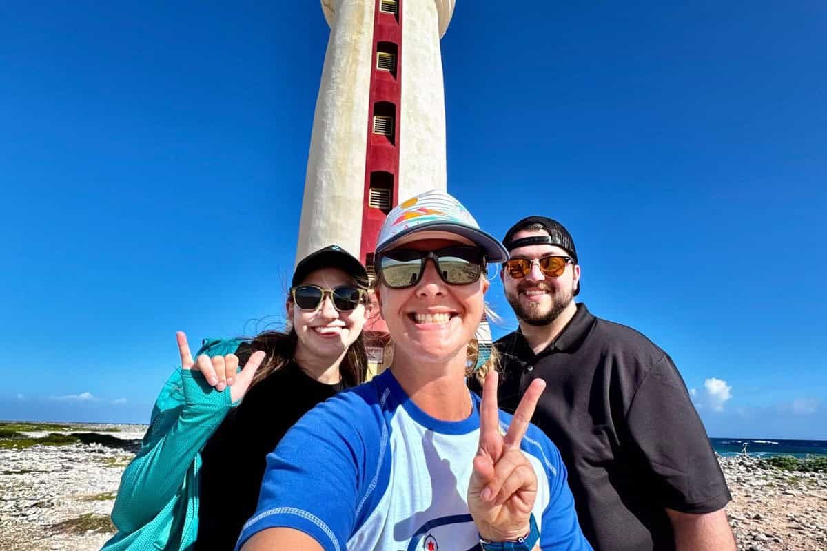 Drie mensen glimlachen en poseren voor een selfie op een zonnige dag tijdens de Privé Zuid Tour (Bonaire), met een hoge vuurtoren en blauwe lucht achter hen. De voorste persoon knippert met vredestekens, terwijl de andere twee een zonnebril dragen en handgebaren maken.