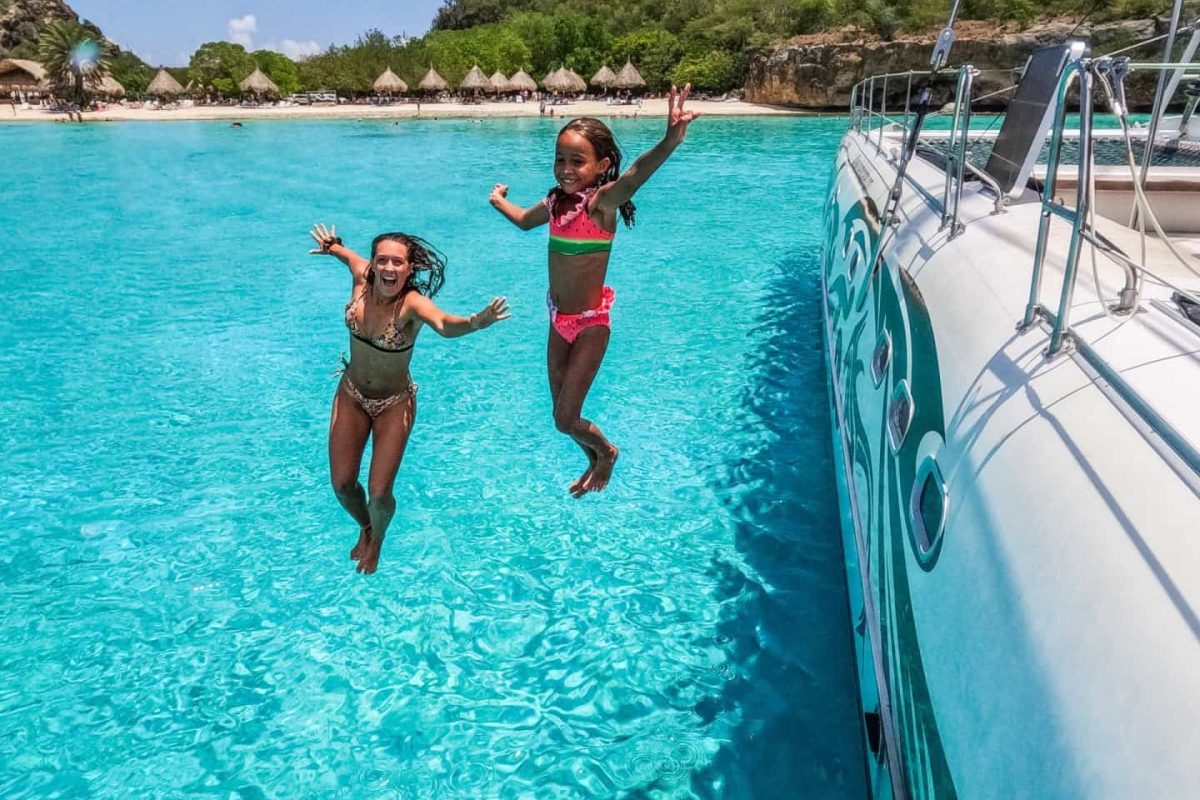 Twee mensen in badkleding springen vrolijk van een boot in het heldere turquoise water tijdens de Strandreis naar de westkust (Curaçao), met palmbomen en groene natuur op de achtergrond.
