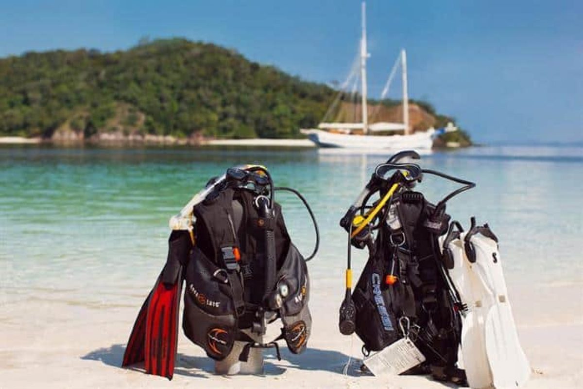 Dos equipos completos de buceo de alquiler, incluyendo equipos de ordenador de buceo (Bonaire) descansando en una playa arenosa, con aletas y tanques, mientras un velero flota cerca de islas verdes y mares tranquilos de fondo.