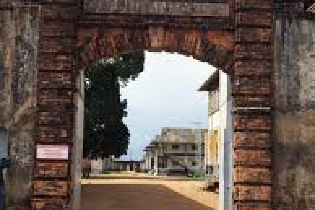 The French Guiana Day Trip (Suriname) has a large weathered brick arch entrance to a compound with historic buildings and a dirt track, under a partially cloudy sky.