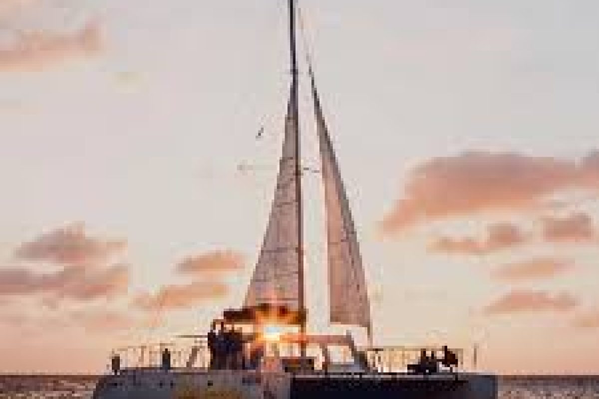 The Sunset Cruise (Aruba) features a catamaran sailboat with passengers gliding on the ocean at sunset, orange and pink clouds in the sky, and sunlight streaming through the sails.