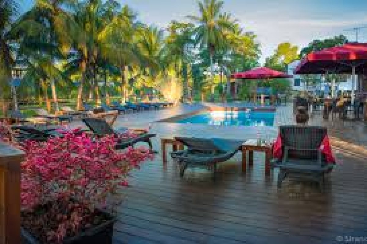 Ervaar de Dagtrip Frederiksdorp (Suriname): lounge bij het zwembad op een houten dek met rode parasols, weelderige palmbomen, roze bloemen en ontspannende zitjes onder een blauwe hemel.