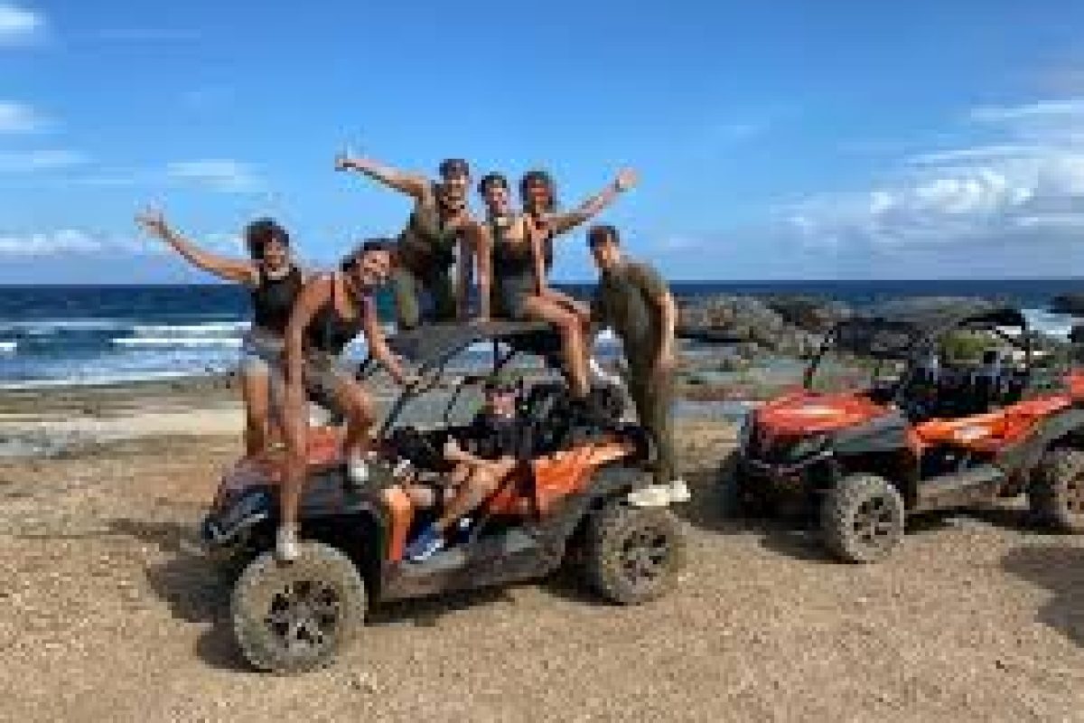 Un grupo de personas sonrientes posa sobre y alrededor de dos vehículos naranjas de ATV Adventure (Curazao) cerca de una playa rocosa, con olas azules del océano y un cielo despejado de fondo.