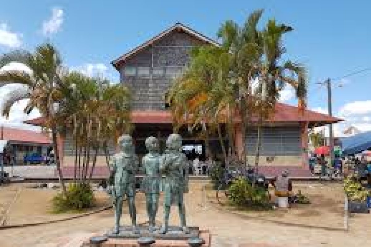 On the French Guiana Day Trip (Suriname), a bronze statue of three children stands in front of a wooden building with a red roof and palm trees, under a blue sky with scattered clouds, with people visible in the background.