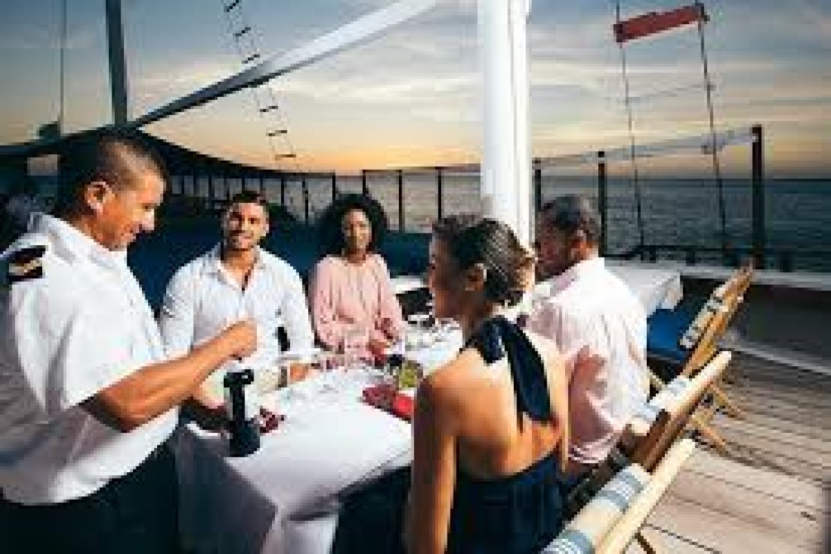 Four guests smile at a dining table on the Sunset Cruise (Aruba) as a uniformed server attends to them. Wine glasses and plates are set, with the ocean and sunset providing a scenic backdrop.