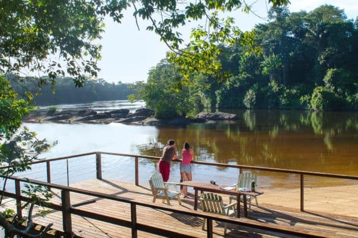 Tres personas están de pie en una terraza de madera junto a un río tranquilo, rodeadas de un denso bosque bajo un cielo despejado, durante el Viaje en Barco de la Sabana Judío (Surinam). Dos sillas vacías y una mesa están en la terraza, con vistas a las rocas en el agua.