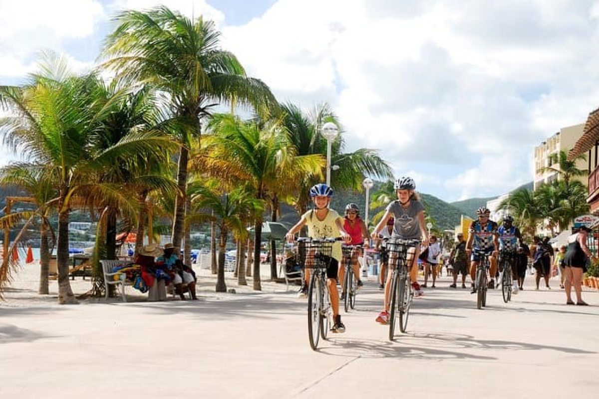Disfruta el Recorrido Histórico en Bicicleta y Tiempo en la Playa (San Martín): recorre con otros ciclistas un paseo junto al mar rodeado de palmeras, con ambiente tropical y personas paseando o descansando a la sombra.