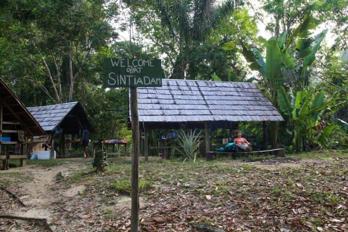 Un cartel de madera con el texto "Bienvenido Opat Sintiadam" se encuentra junto a tejados de paja en un frondoso bosque, donde la gente se reúne durante la Expedición de los 6 Días Gran Río Sintiadam (Surinam).