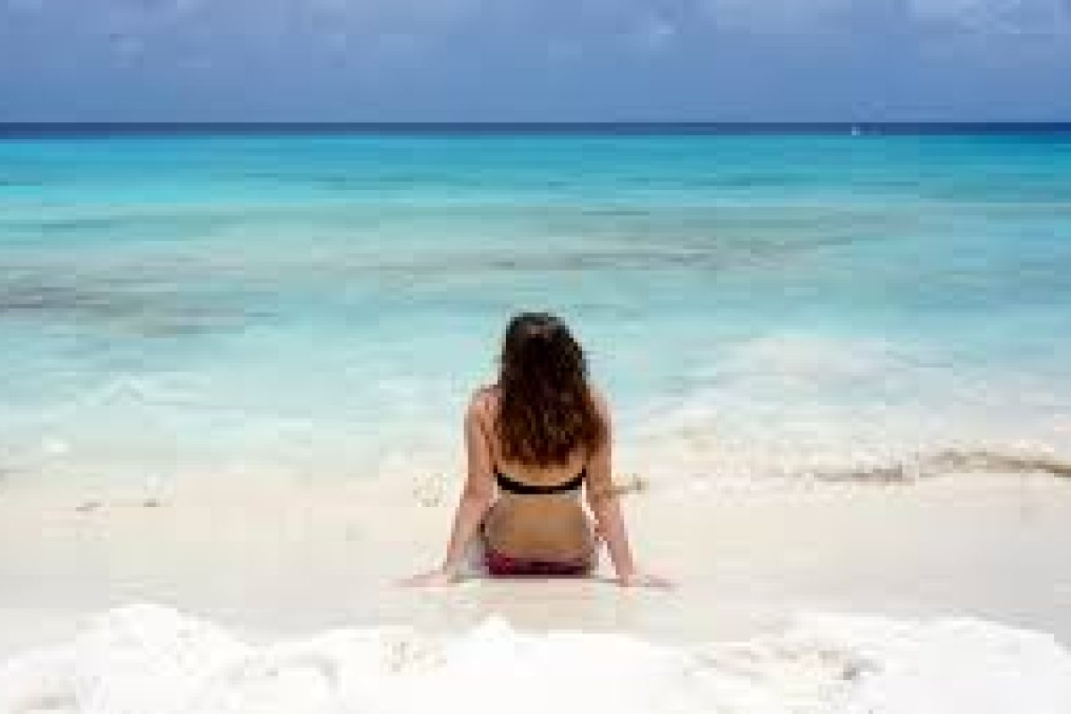 A woman in bikini relaxes on white sand, staring at calm turquoise water under a cloudy sky—a tranquil scene reminiscent of the Klein Curacao Sunset Cruise (Curaçao).