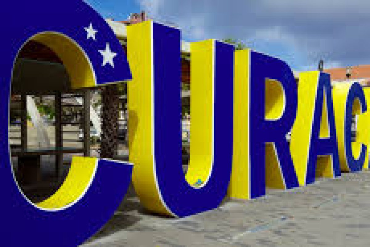 Grote, kleurrijke 3D-letters die "CURAÇAO" spellen staan buiten - blauwe C met twee witte sterren, gele letters met blauwe contouren - tegen gebouwen en palmbomen. Ervaar dit uitzicht tijdens de Eiland Tour (Curaçao).