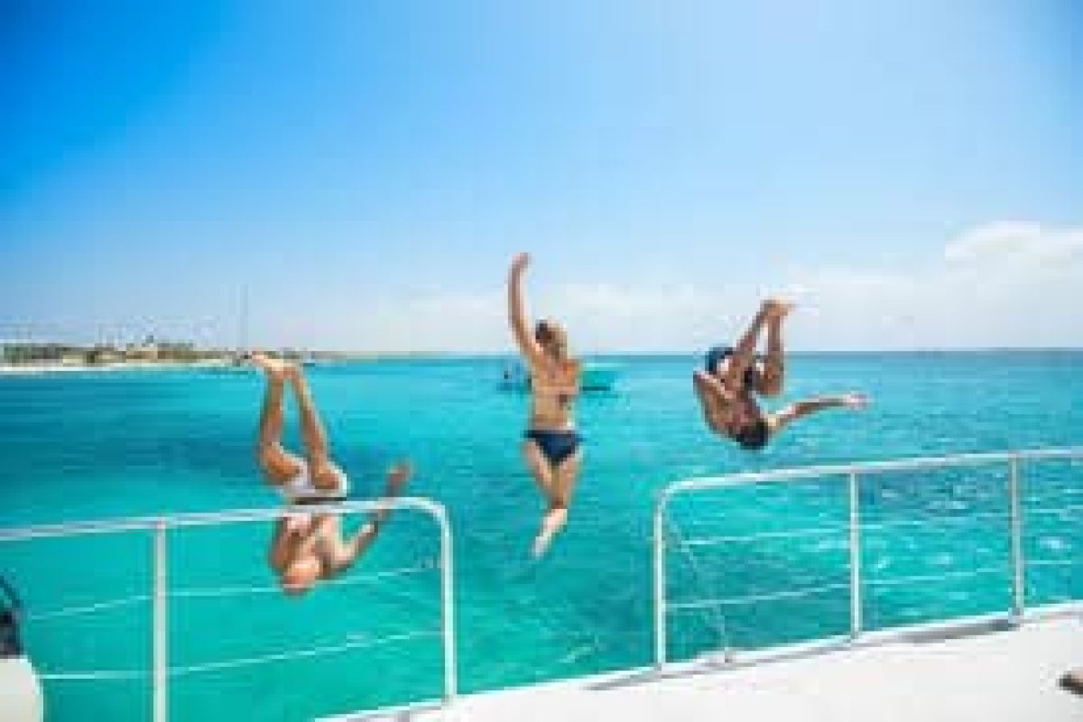 Tres personas saltan al agua turquesa desde un barco durante el Crucero de lujo por la laguna (Aruba), bajo un cielo azul y con la costa a lo lejos.