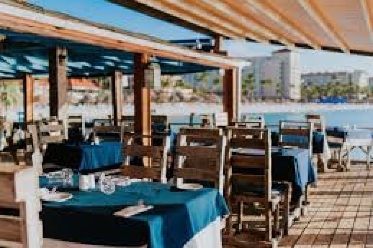 Disfruta de un crucero al atardecer con cena de cuatro platos (Aruba): cena al aire libre en mesas sombreadas con ropa de cama azul y blanca, cristalería y cubiertos mientras recorres el soleado malecón con vistas a los edificios a lo lejos.