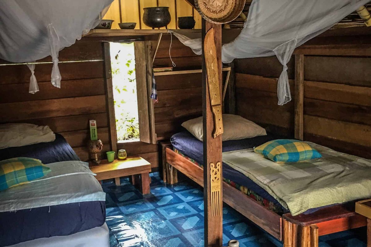 The 3 Days Galibi Myrysji (Suriname) has a rustic wooden cabin interior with two single beds draped in white mosquito nets, a blue-patterned floor, a central wooden pole, wall baskets and a large clay jug on the floor.
