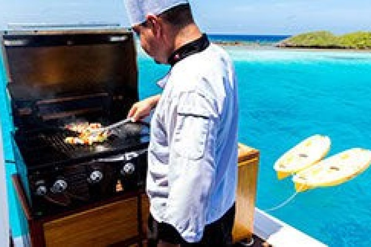 Durante el Crucero de lujo por la laguna (Aruba), un chef vestido de blanco asa comida en una barbacoa instalada en un barco rodeado de agua azul brillante y pequeñas islas, con dos flotadores amarillos cerca.