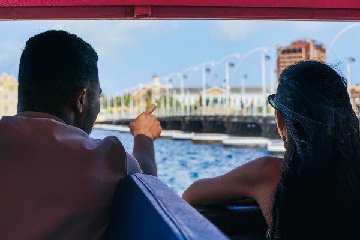 Twee mensen op de boot van Eiland Tour (Curaçao) kijken naar een waterkant. De ene wijst naar een brug en kleurrijke gebouwen onder een blauwe lucht. Het tafereel is ingekaderd vanuit de boot.