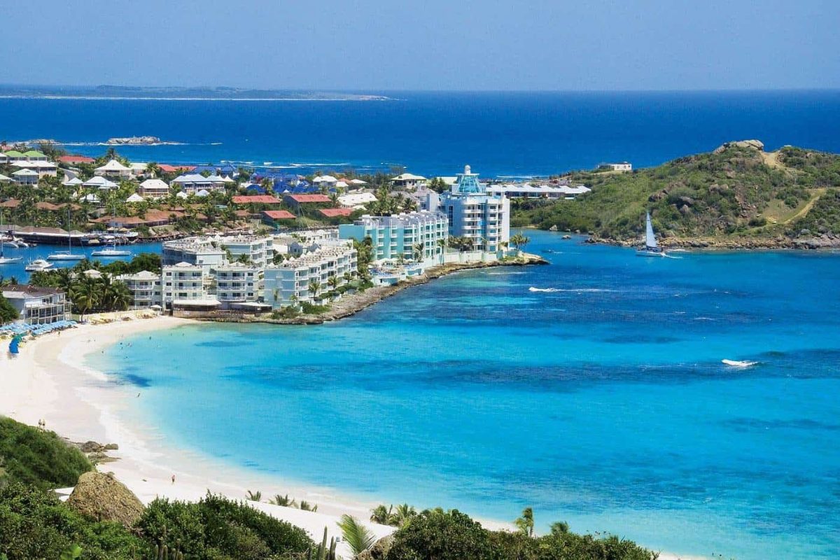 Bekijk de kustlijn van Sint Maarten vanuit de lucht met turquoise water, zandstranden, witte gebouwen, groen en een zeilboot onder een blauwe hemel - ervaar het allemaal tijdens de Gem of the Island Tour met Ebike.