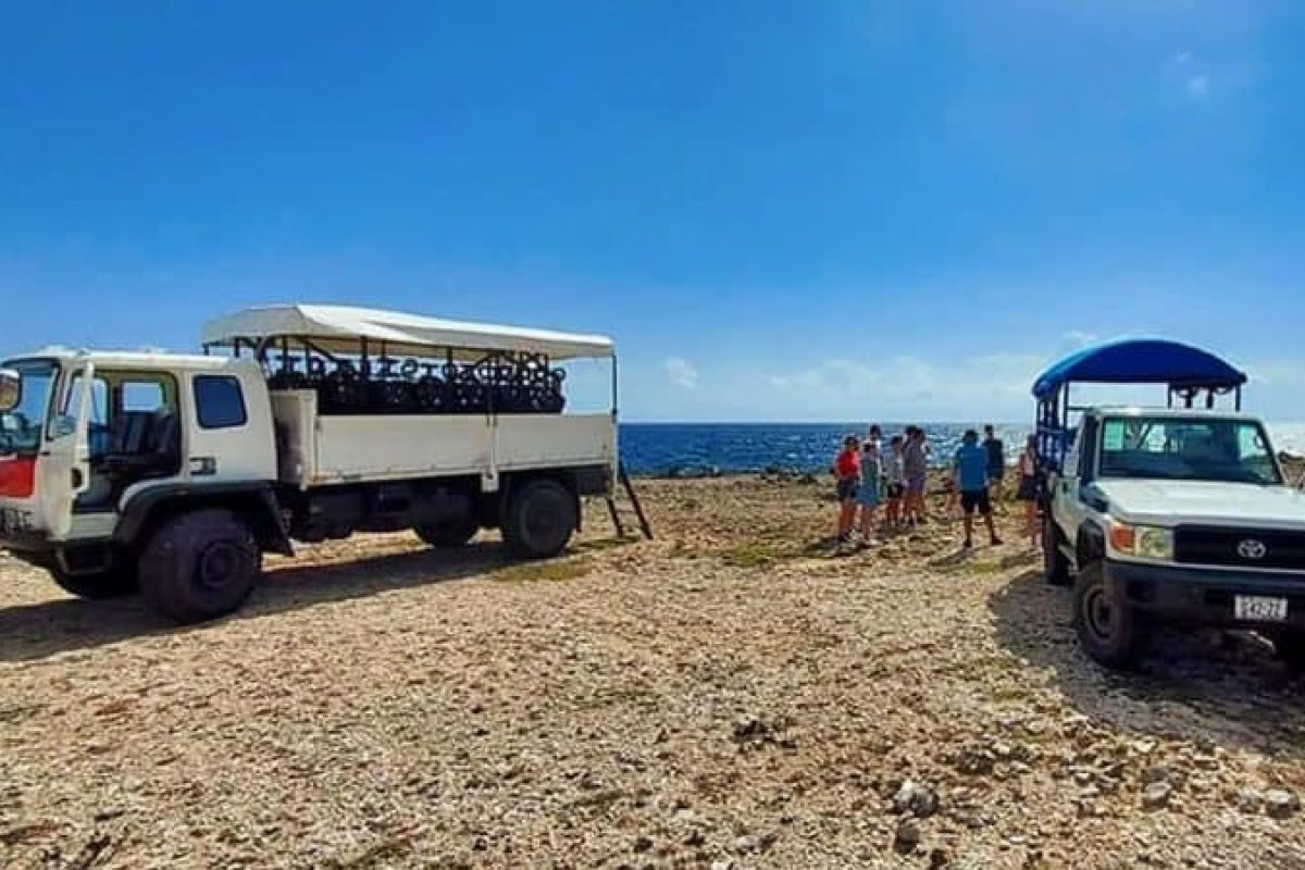 Twee vrachtwagens staan geparkeerd op een rotsachtige kust in Curaçao onder een blauwe lucht. Een groep bereidt zich voor op Snorkel en strandavontuur, met duikflessen zichtbaar op de ene truck.