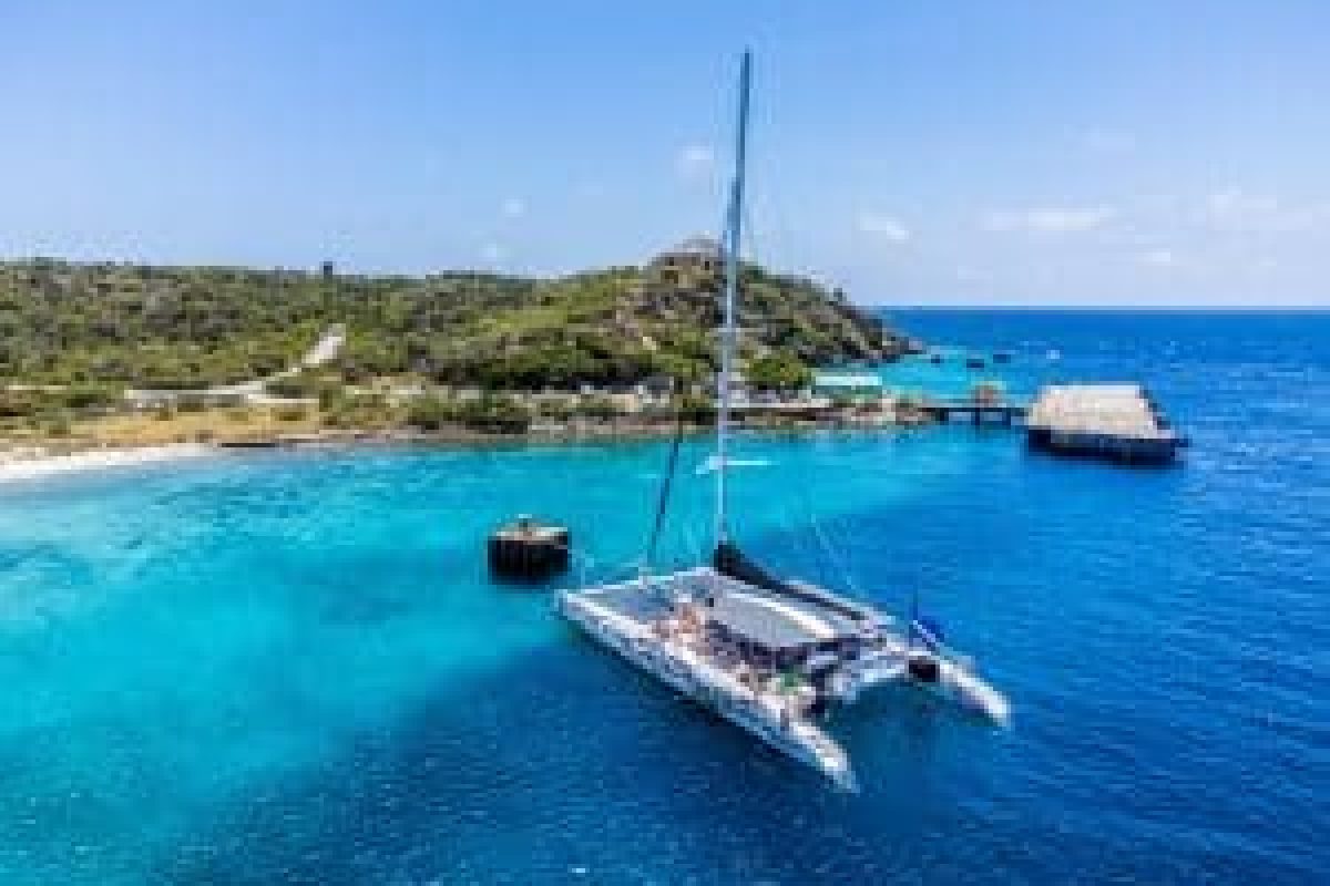 The Klein Curacao Sunset Cruise (Curaçao) features a spacious catamaran that glides over turquoise waters near a lush sandy island with a pier, offering spectacular views under clear skies for guests on board.