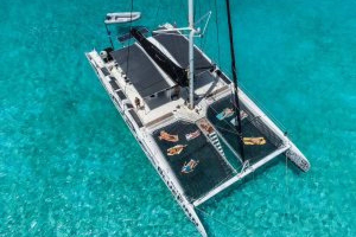 Vista aérea de un catamarán blanco del 3 en 1 Snorktocht (Curazao) con personas relajándose sobre redes sobre aguas turquesas claras. Un pequeño bote está enganchado a la parte trasera y el mar tranquilo revela el fondo arenoso poco profundo.