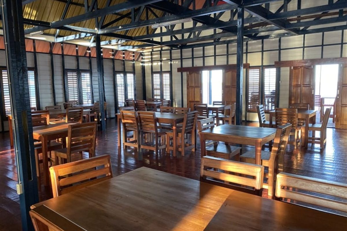 Experience the 3 Days Bigi Pan Akira Overwater Resort in Suriname, featuring a spacious, sun-drenched dining area with wooden tables and chairs, high ceilings, large windows with wooden shutters and polished wooden floors for a warm ambiance.