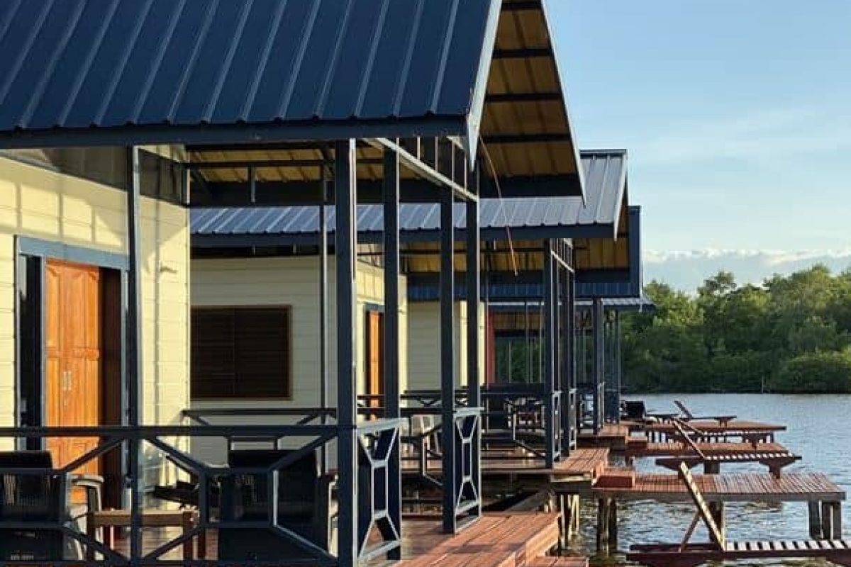 At the 3 Days Bigi Pan Akira Overwater Resort (Suriname), wooden cabins with blue metal roofs stand on stilts above the water, each with a deck with tables and chairs, against trees and a clear sky in the background.